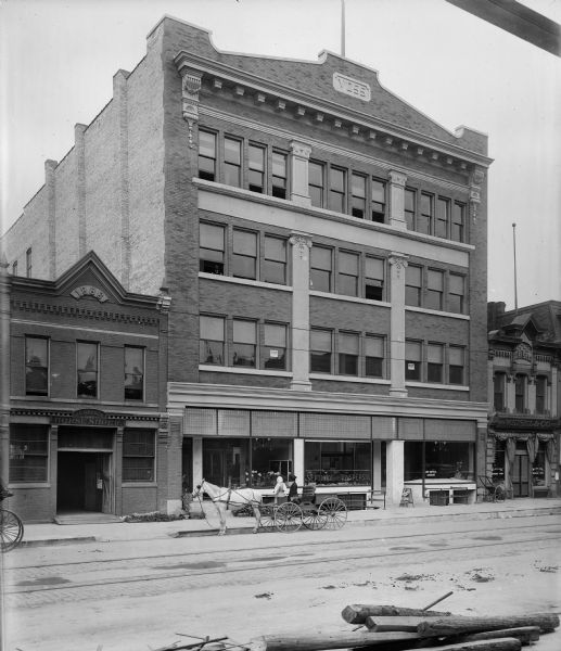 The Voss Building on N. Water Street between E. Juneau Avenue and E. Highland Avenue. J.C. Grosch Horse Shoer is to the left. There is a horse-drawn vehicle with two women parked in front of the Voss building.