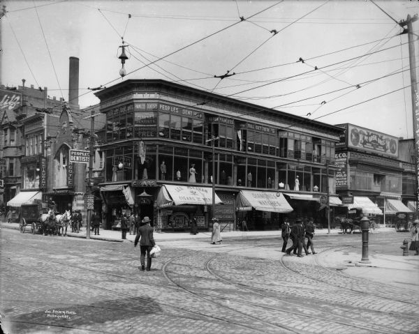 Street corner with commercial buildings, including Leo Abraham Cigars and Merit, Gargoyle Restaurant, The Peoples Dentists, The Hermit Buffet, and surrounding commercial buildings. Pedestrians in foreground are walking in the street, along with horse-drawn vehicles.