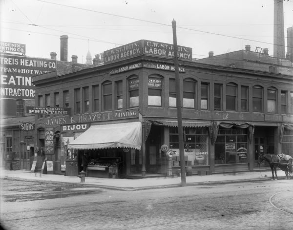 Northeast corner of N. 2nd and W. Michigan streets with the James G. Brazell Printing and C.W. St. John Labor Agency businesses. A man is standing at a fruit and vegetable stand, there are signs for Pabst, and a scale is outside of a storefront advertising "Fine Liquors Cigars," and "Wegner Bros. Steam & Hot Water Heating."
