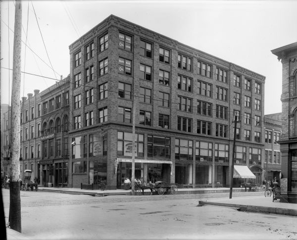 Watkins building at the southeast corner of N. 3rd Street and W. Kilbourn Avenue. Businesses include: Reimers Photo materials, Provident Loan Society, Arthur Koenig Company, and Leo. Vogel & Co. Tailors. On the opposite corner is C.J. Artist, Tailor. On the extreme right can be seen the Williams & Brenckle Cigar Mfg. Co., with a wooden Indian in front. There are horse-drawn vehicles parked on the curb on the street.