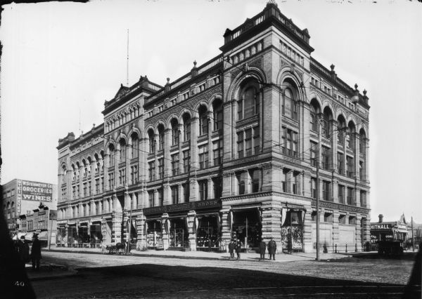 Metropolitan building and the Streissguth clothing and shoe company at the northeast corner of N. 3rd and W. State Streets. Police officers are standing on the street corner among other men and parked, horse-drawn vehicles.