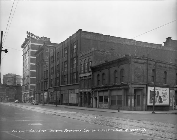 Looking northeast from N. Water and E. Buffalo Streets. Cars are parked on the side of Water Street, and streetcar tracks run down the street. Caption on negative reads: "Looking northeast showing property side of street — 342 N. Water St." Two businesses are the Marber Paper Co., and Resnick's Handbag Co. A large sign (in reverse) on top of a roof building reads: "Mayer-Hughes:.