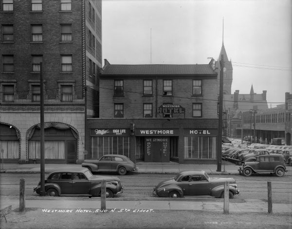 The Westmore Hotel, located at N. 5th Street near W. Clybourn Street. Automobiles are parked on both sides of the street and in a parking lot to the right of the hotel. In addition, the Milwaukee Road Depot is in the background. Caption on print reads: "Westmore Hotel, 540 N. 5th Street".