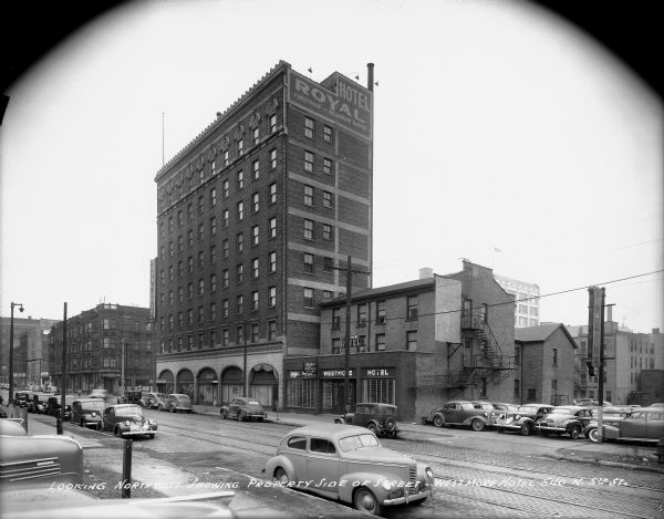 Westmore Hotel and surrounding commercial area looking north from N. 5th and W. Clybourn Streets. Automobiles parked on both sides of the street and in parking lot. Caption on print reads: "Looking northwest showing property side of street — Westmore Hotel 540 N. 5th St".