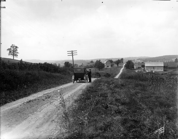 Man, possibly Joseph Brown, attending to an automobile on the road between Waukesha and Ionsman. Rural landscape and farm in the background.