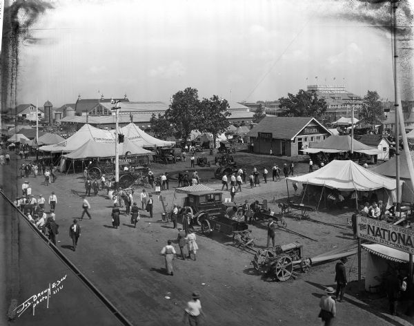 Numerous tents with farm machinery and technology with the colosseum in the background, presumably the Wisconsin State Fair.