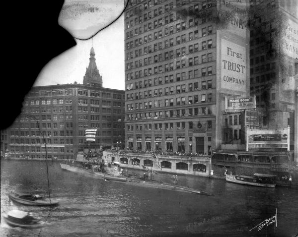Elevated view up the Milwaukee River from Wisconsin Avenue bridge. A World War I submarine travels down the Milwaukee River. A crowd is gathered at the riverbank, and individuals are looking out of the First National Bank windows.