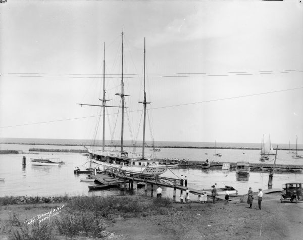 Lily E., a schooner converted to the South Shore Yacht Club's floating club house, until the winter of 1921. The club house is at the foot of Nock Street in a Bay View neighborhood. Numerous other sailing and paddle boats surround the yacht while people on shore and sitting on the pier look on. Lake Michigan is in the background.
