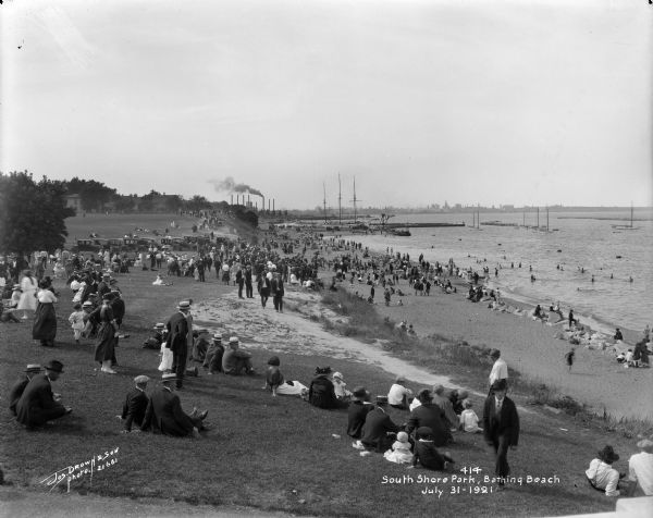 Crowds of individuals along South Shore Beach in the Bay View neighborhood. Some individuals are swimming while others are sitting on the grass and sand.  Metropolitan area in background. Caption on negative reads, "South Shore Park, Bathing Beach".