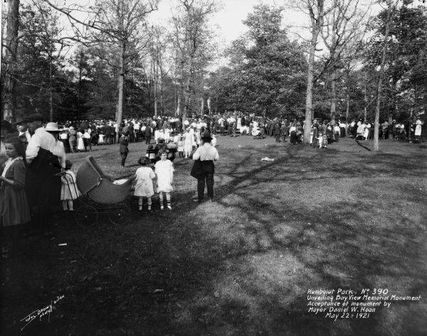 A crowd of people at Humboldt Park gather for the unveiling of a World War I monument. A family in the foreground with larger crowd in the background. Caption on negative reads, "Humboldt Park. Unveiling Bay View Memorial Monument. Acceptance of monument by Mayor Daniel W. Hoan".