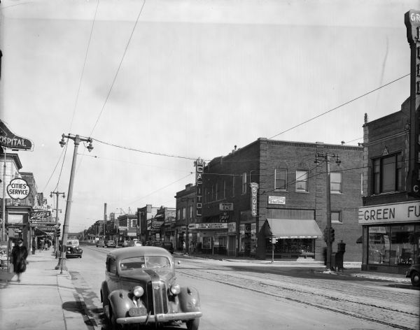 Looking west from S. 73rd Street at the Capitol Theatre and surrounding commercial buildings along W. Greenfield Avenue. Automobiles are parked on both sides of the street, and pedestrians are walking on the sidewalk in front of the commercial buildings.