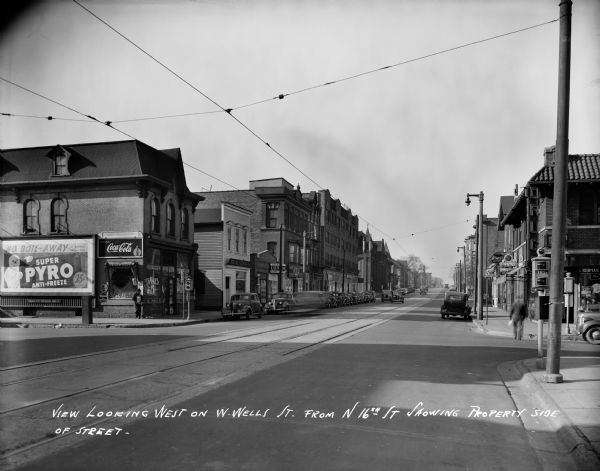 Looking west on W. Wells Street from N. 16th Street. Automobiles are parked on both sides of the street and  a large billboard advertising is in the left foreground. Caption on negative reads: "View Looking West on W. Wells St. from N 16th Showing Property Side of Street."