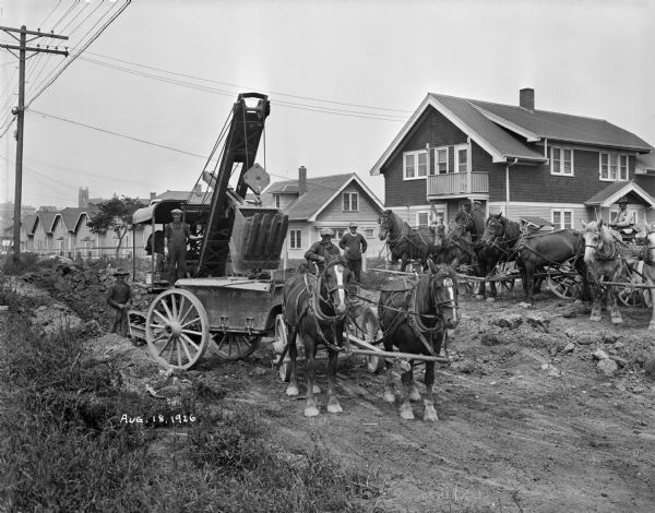 Construction workers and early construction machinery being driven by horses in a residential neighborhood.