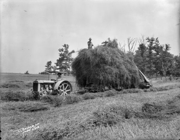 Three men harvesting wheat. One man is driving a tractor, while the other two men are on top of the hay loaded onto a wagon.