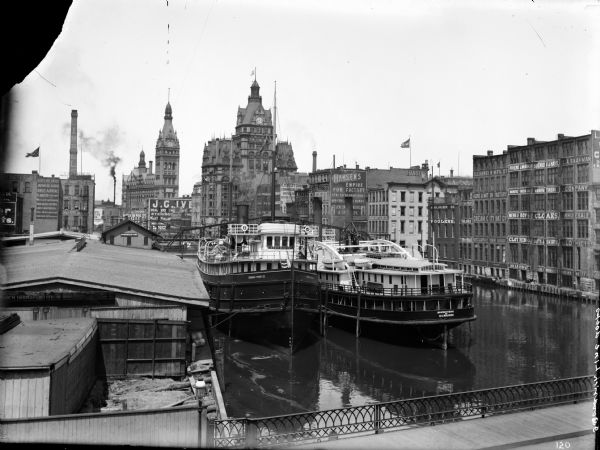 Elevated view of two Goodrich Line passenger ferries docked on the west bank of the Milwaukee River, north of the Clybourn Street bridge. The Pabst building and City Hall are in the background, with other commercial and manufacturing buildings.