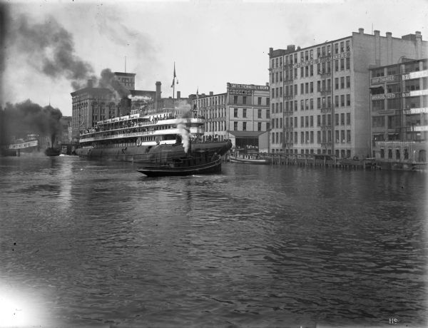 Looking north on the Milwaukee River as the whaleback excursion steamer "Christopher Columbus" is guided by tugboats at about St. Paul Avenue.