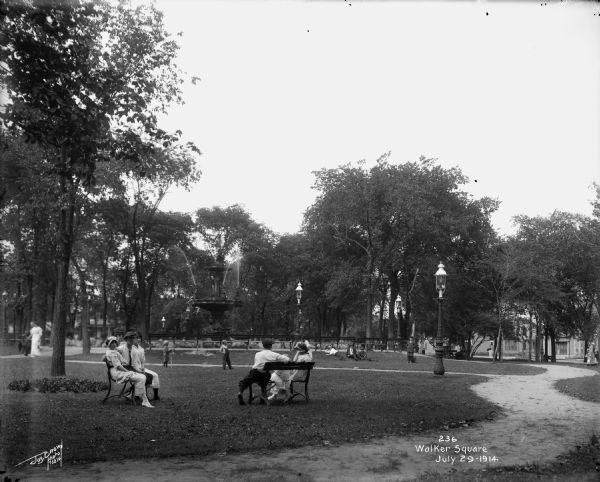 Individuals and children sitting on benches in the foreground of the residential park, with others in the middleground laying on the grass near the water fountain. Caption on glass plate reads: "Walker Square".