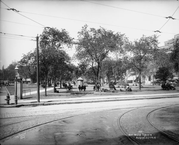 View from across the street of 4th Ward Square. Individuals are sitting on benches and on the grass in the park. Caption on glass plate reads: "4th Ward Square".