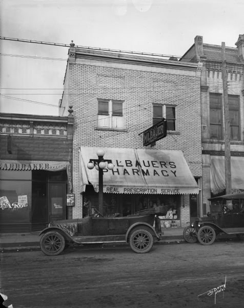 Storefront view of Milbauer's Pharmacy, with two automobiles parked on the street.