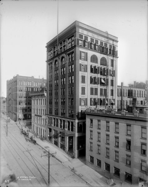 Elevated view of the south side of Mason Street looking east from N. Water Street, featuring the Sentinel Building. There are open windows in the Sentinel Building which has decorative awnings. Several horse-drawn vehicles are parked in front of the Sentinel Building.