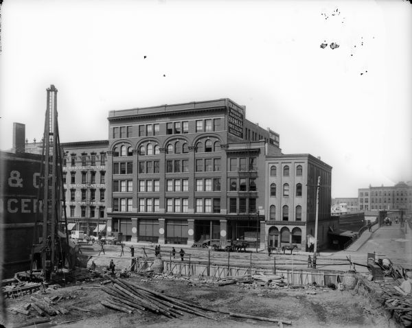 Elevated view of west side of N. Water Street between E. Buffalo and E. Chicago Streets, featuring the Joys Building, with a contruction area in foreground. There are also construction workers, raw lumber and machinery present.