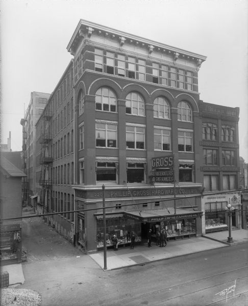 Elevated view of the storefront of the Philip Gross Hardware Company on the east side of N. Third Street, north of W. Wells Street. Grassler and Gezelschap is next door on the right. Part of an alley is on the left.