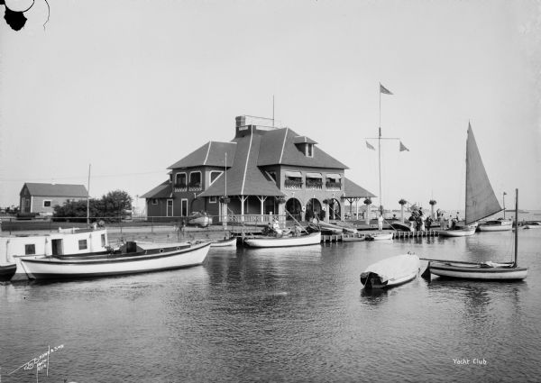 Milwaukee Yacht Club, located at the foot of E. Layfayette Street on Lake Michigan. Numerous sailboats are in the harbor, and individuals are on the pier.