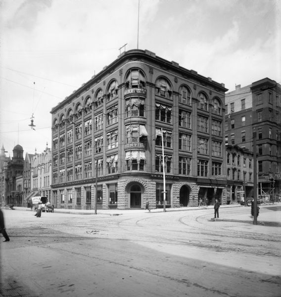 View from across the street of the Hathaway building, probably at the northeast corner of E. Mason Street and N. Broadway. W.M. Marnitz Co. Tailors, Gilbert Commercial College, and other unidentified businesses are located inside the Hathaway building. Several pedestrians are walking on the sidewalks and streets.