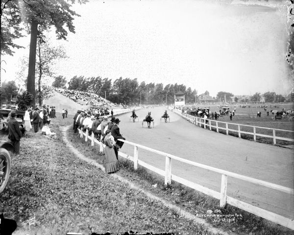 Crowds at the race course watching harness racing in Washington Park. There are three racers, and a crowd of spectators is sitting in the grandstands, on the fences, and standing in the grass areas.