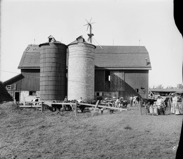 Barn, silos and cows at a farm. There are men attending to the cows, while the women and children are attending to the milk cans.