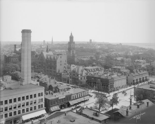 Elevated view looking northeast from the roof of the Wells Building at E. Wisconsin Avenue and N. Milwaukee Street. St. John's Cathedral is in the center of the photograph, surrounded by various commercial buildings and a residential neighborhood, with the shoreline of Lake Michigan in the background.
