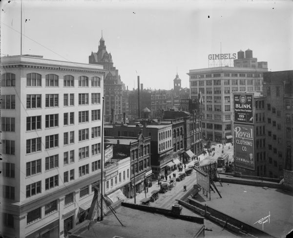 Looking southeast from the roof of the Germania Building, N. Plankinton Avenue and W. Wells Street. Automobiles are parked along the curb, and pedestrians are walking on the sidewalks. There is a Gimbels sign on a building in the background.