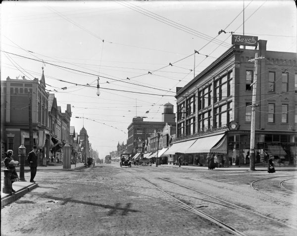 Looking south on N. 3rd Street from W. North Avenue. Bauch goods and other unidentified businesses visible on 3rd Street. There are automobiles and streetcars driving on the street, and several pedestrians on the sidewalks and street.