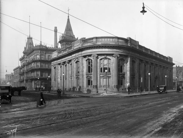 Second Ward Savings Bank building (now the Milwaukee County Historical Society), looking northwest from the corner of W. Kilbourn Avenue and N. Plankinton Avenue. The Republican Hotel is in the background at 343 Third Street. In addition there are automobiles, horse-drawn vehicles, and pedestrians. This bank building was designed by the architectural firm of Kirchhoff and Rose. Listed on the National Register of Historic Places, it was originally constructed in 1913 as the Second Ward Savings Bank. The Milwaukee County Historical Society moved into the building in 1965, and opened its first exhibition the following year.