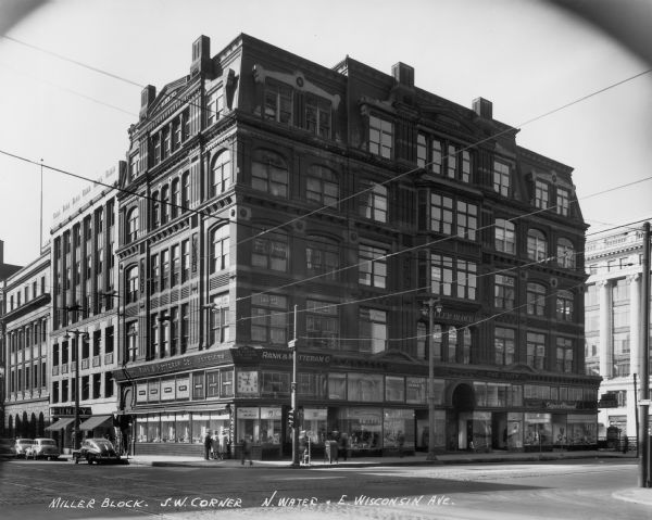 Mack building on the southwest corner of N. Water Street and E. Wisconsin Avenue. Rank and Motteram Co. jewelry business inside the Mack building. There are several pedestrians on the sidewalk, and automobiles parked at the curb.