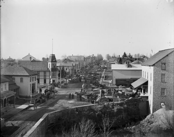 Elevated view of many horse-drawn vehicles on a main street in an unidentified rural village, perhaps at a farmer's fair. A crowd of men is standing outside a hotel on the left side, and a railroad car with "Chicago, Milwaukee & St. Paul" written on it is sitting on tracks near commercial buildings on right side of image.