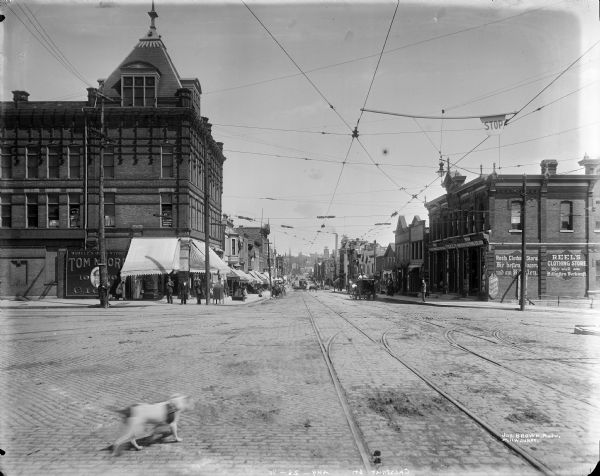 W. Juneau Avenue commercial district looking east from N. 7th Street. A dog is crossing the street in the foreground, and pedestrians, and horse-drawn vehicles are in the distance.