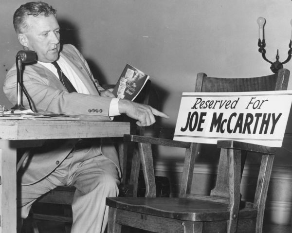 Leonard Schmitt, independent Republican candidate for the United States Senate, holding a copy of <i>The McCarthy Record</i> and pointing to an empty chair with a sign showing the seat reserved for his opponent, Joseph R. McCarthy, to debate the issues with him. 
