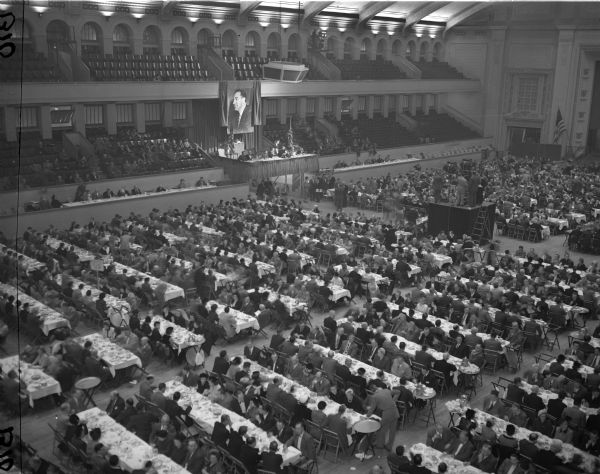 Elevated view of a crowd gathered for a dinner that honored Senator Joseph R. McCarthy. Behind the elevated head table is a large portrait of the Senator.
