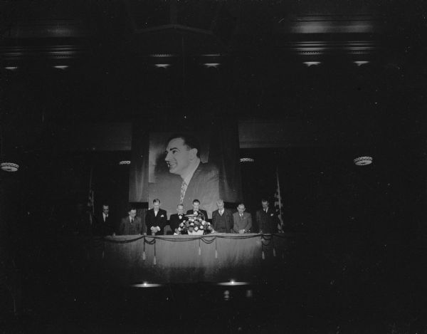 Head table at a dinner that honored Senator Joseph R. McCarthy, behind which is a large portrait of him. Senator McCarthy is second from the right. Over 2500 Wisconsin Republicans paid $25 each to hear McCarthy launch his campaign for re-election. The senator was reportedly so overcome by the cheers he received that he was barely able to speak.