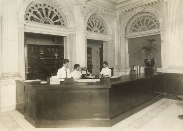 The Historical Society Library delivery desk and librarians Mary S. Foster, Esther DeBoos, and Edna C. Adams, 1912.  At the time, patrons and students were not admitted to the stacks; instead they filled out call slips and the books were paged and shelved until pickup in the mahogany bookcases behind the women.