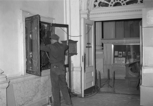 Electrician feeding cable into the electrical box on the north second floor hallway of th Wisconsin Historical Society. Changes in the library delivery area and lettering on the library doors can be seen behind him.