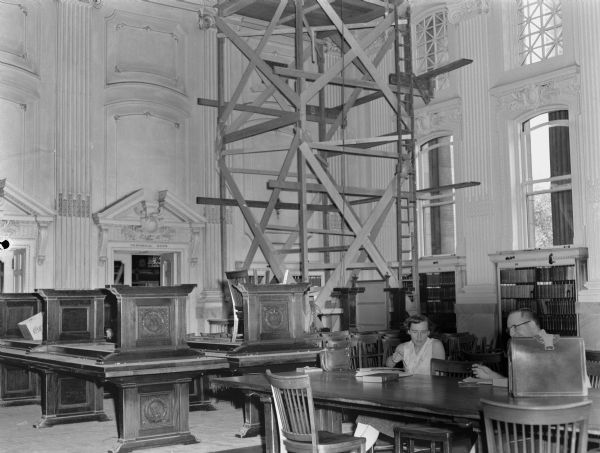 Ceiling high scaffolding in the Wisconsin Historical Society Library reading room, with a man and a woman attempting to study at the tables in the foreground.  The scaffolding was used to remove the original glass skylights and ceiling light fixtures.