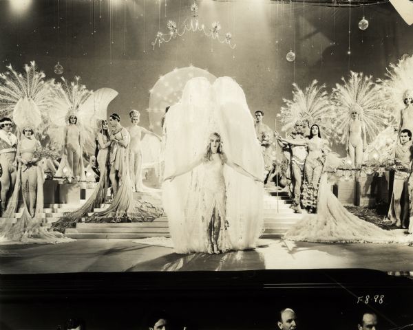 Dancers in elaborate costumes and huge headdresses perform onstage in "Glorifying the American Girl," a 1929 Paramount musical. The heads of some pit musicians are visible in the foreground.