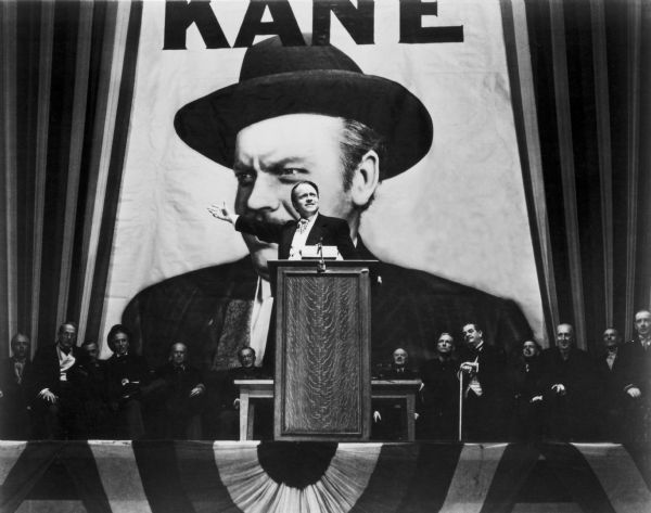 Orson Welles on podium in front of giant 'Kane' backdrop, in scene still for "Citizen Kane".