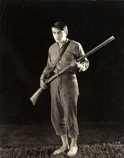 Full-length studio publicity still of Richard Barthelmess as David Kinemon in <i>Tol'able David</i> (First National 1921). He is barefoot, wears rough clothing, and carries an old hunting rifle.