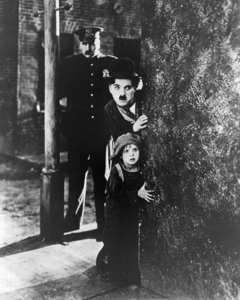 The tramp (played by Charlie Chaplin) and the kid (Jackie Coogan) peer around a wall with the police officer (Tom Wilson) standing behind them, from <i>The Kid</i> (First National 1921).