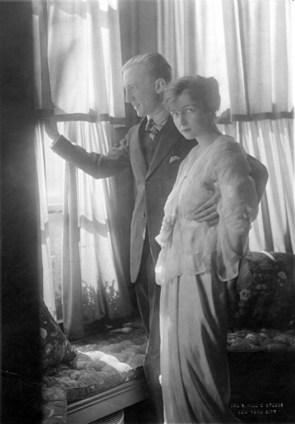 Attractive portrait of the husband-wife dance team, Vernon and Irene Castle standing in a bay window photographed by Ira L. Hill's Studio of New York.