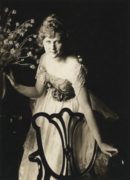Studio portrait of Mary MacLaren, Bluebird Photoplays star, in a formal dress leaning on a chair.