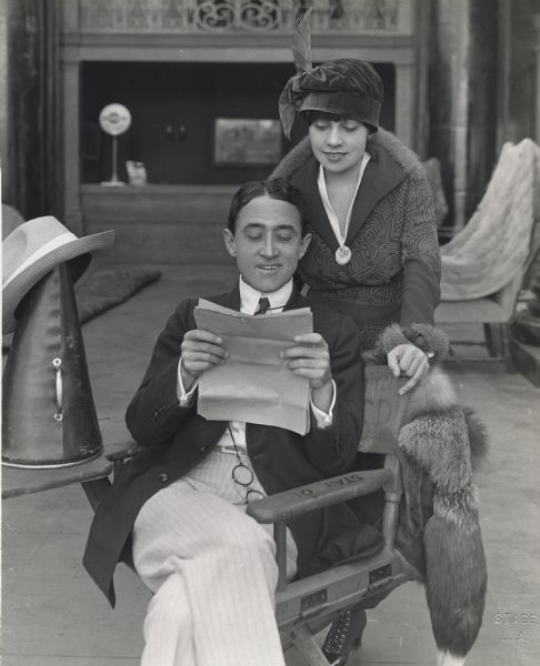 The writer Anita Loos and the director John Emerson looking over a script on a film set. Emerson has a pince-nez on a cord and sits in a director's chair. His hat is perched on a megaphone.